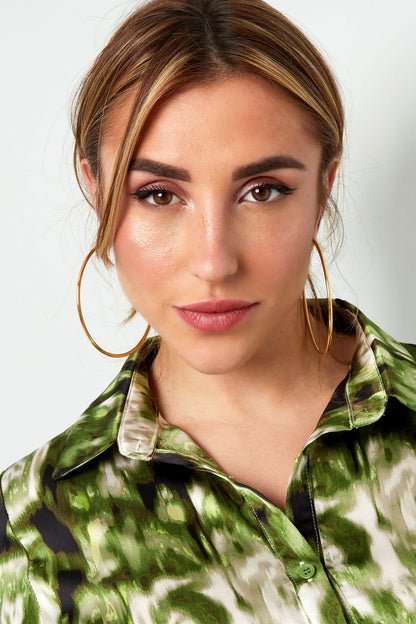 Model wearing large polished gold hoop earrings paired with green patterned blouse, smiling at camera