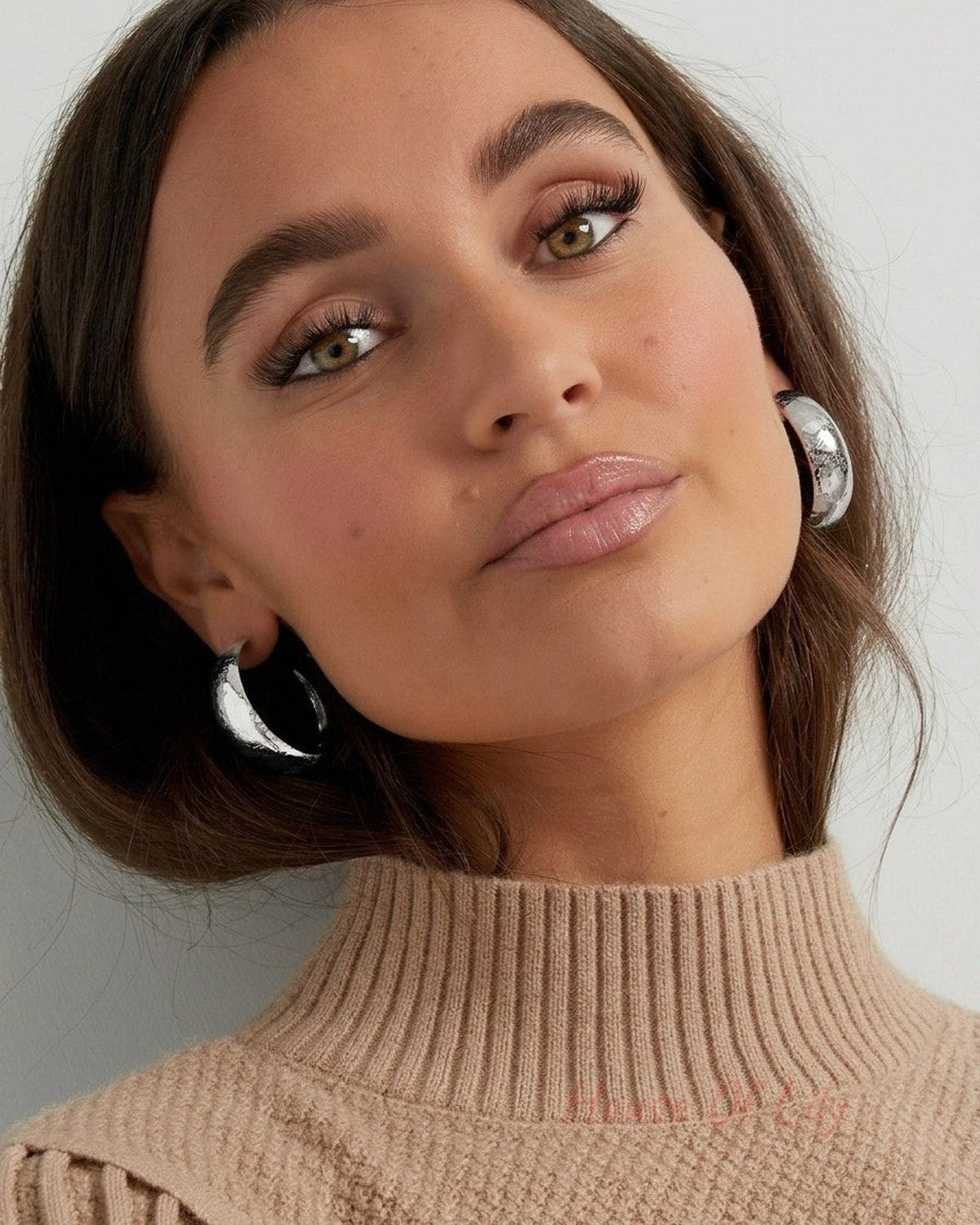 Thick, silver-finished hammered hoop earrings worn on earlobes.