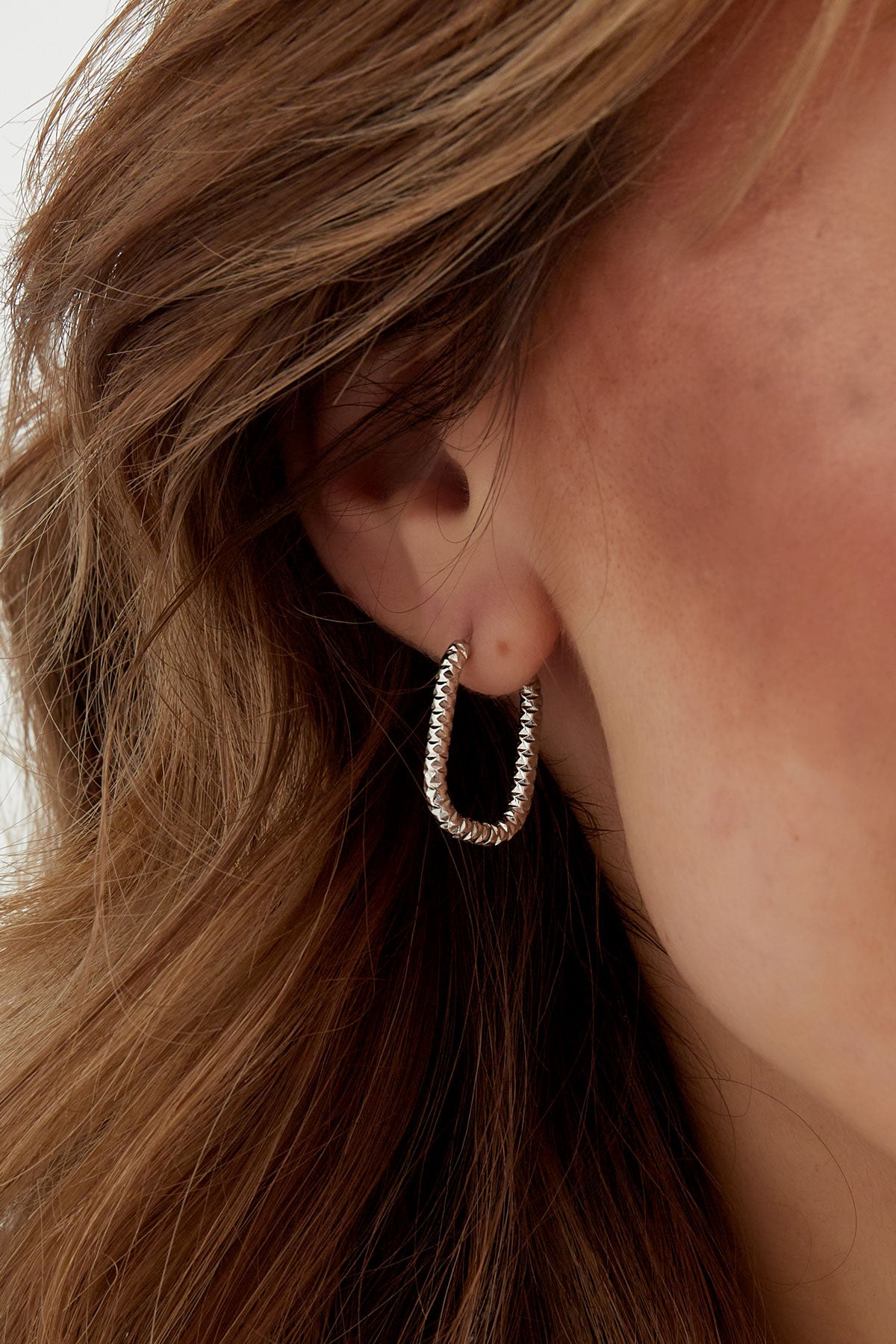 Side profile of model showing silver textured oval hoop earrings with wavy hair