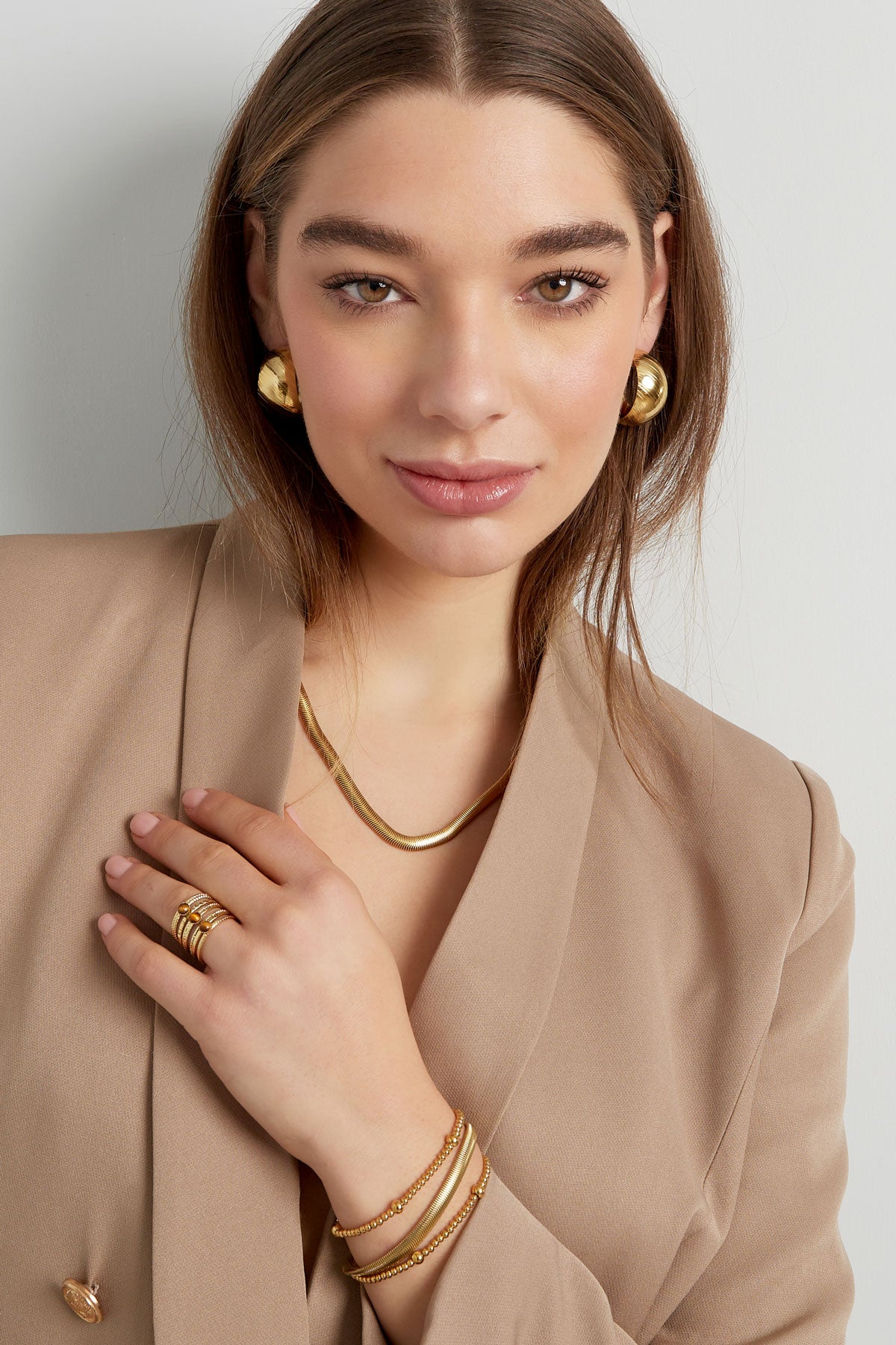 Woman wearing gold chain bracelet, gold necklace, matching ring, and earrings with a beige blazer