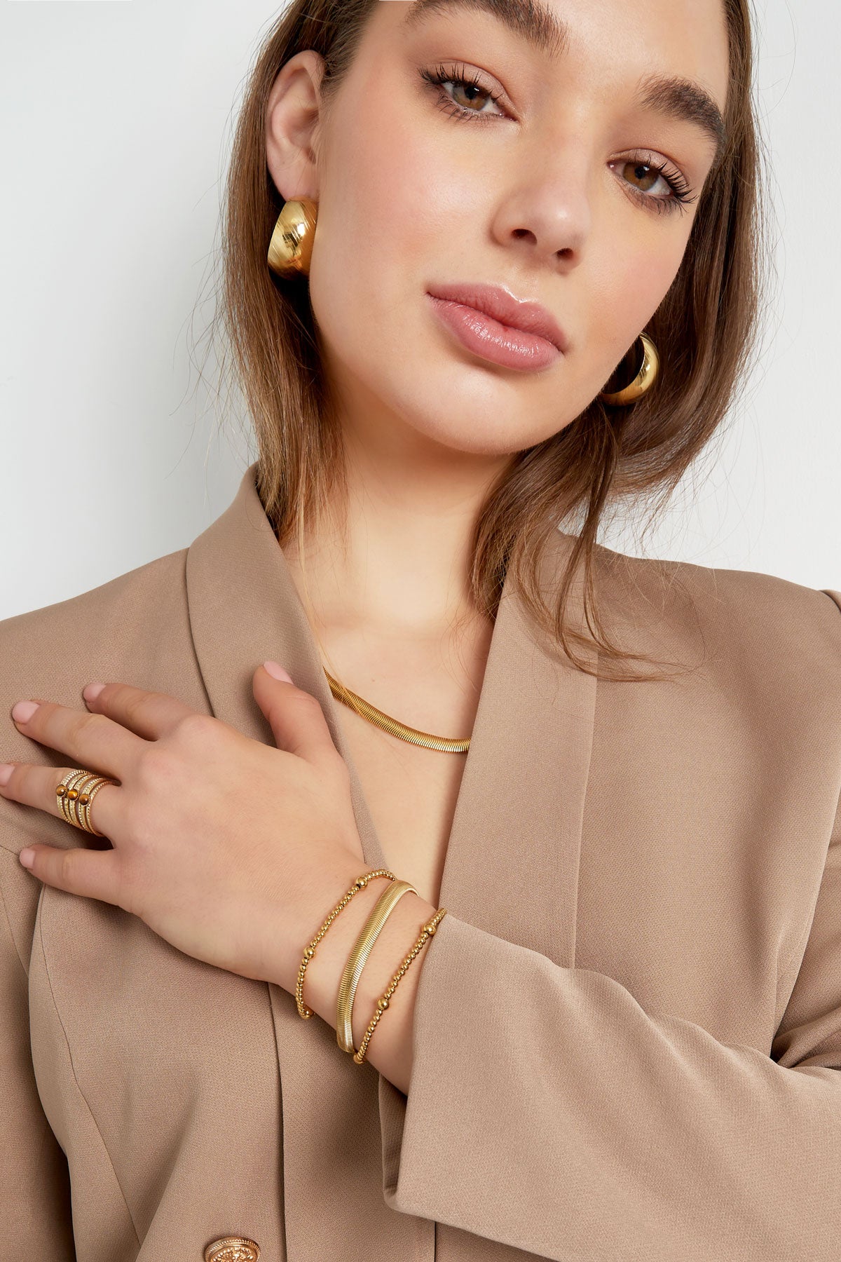 Woman showcasing gold chain bracelet along with matching ring and earrings in beige blazer