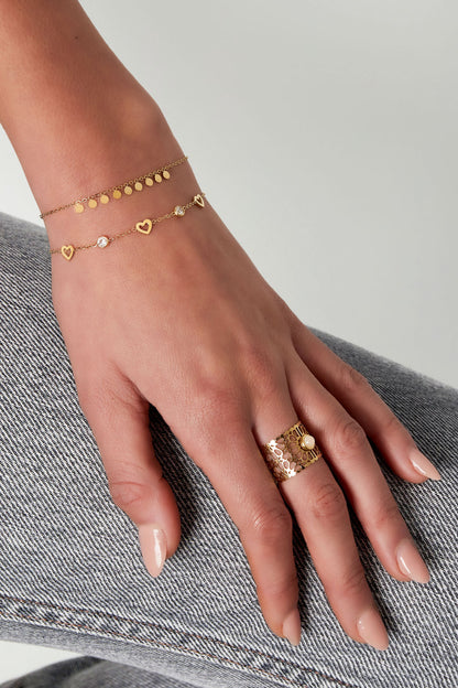 Wrist wearing two gold chain bracelets including one with heart charms and rhinestones, paired with a decorative gold ring and light nail polish
