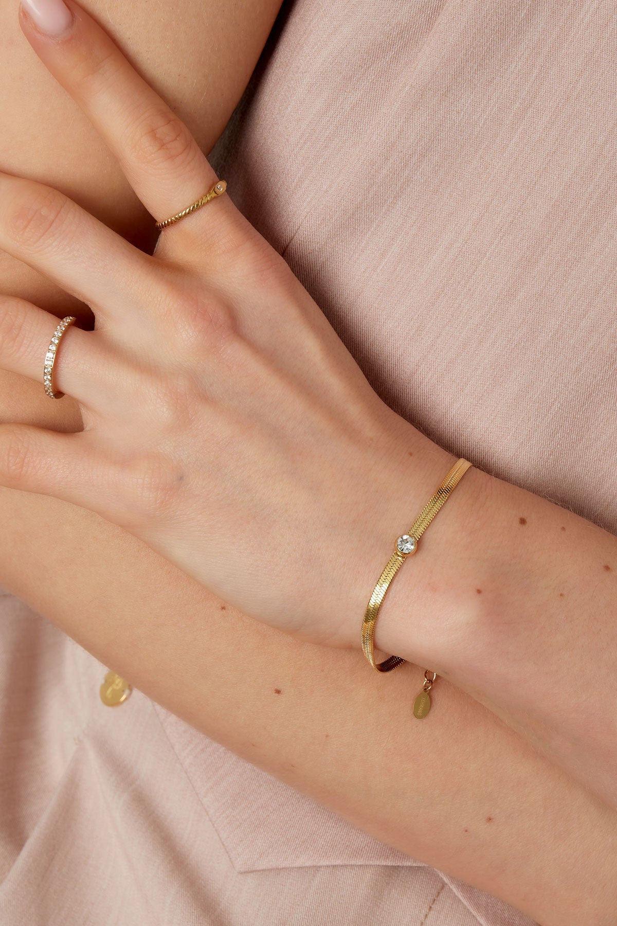 Woman's wrist wearing a slim gold chain bracelet with a sparkling crystal, shown with delicate gold rings against a soft beige top