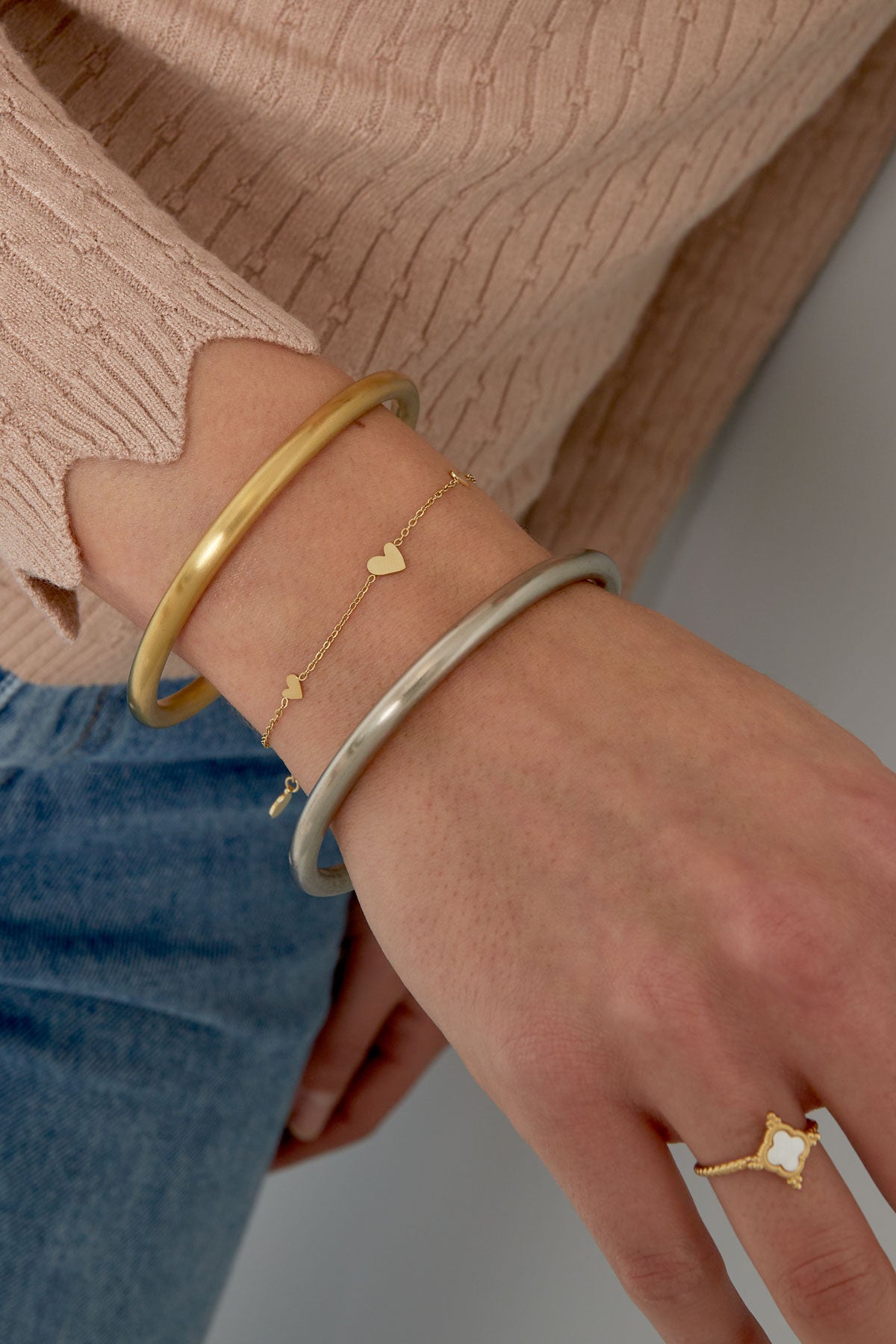 Close-up of a woman's wrist wearing the delicate gold heart charm bracelet alongside two bangles, styled with beige sweater and blue jeans