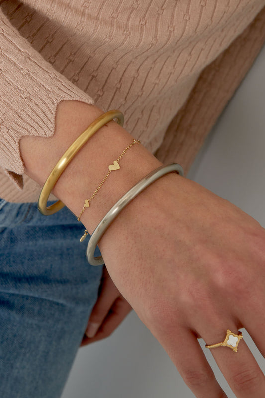 Close-up of a woman's wrist wearing the delicate gold heart charm bracelet alongside two bangles, styled with beige sweater and blue jeans