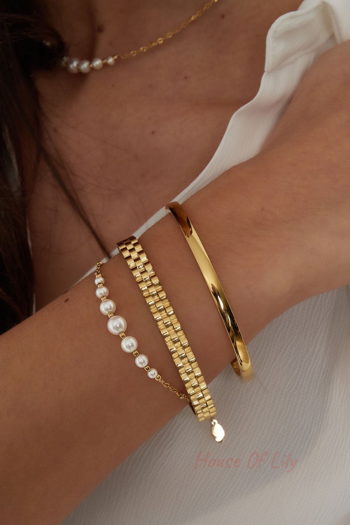 Model wrist wearing layered gold jewelry including a polished gold bangle, gold chain bracelets, and pearl chain necklace
