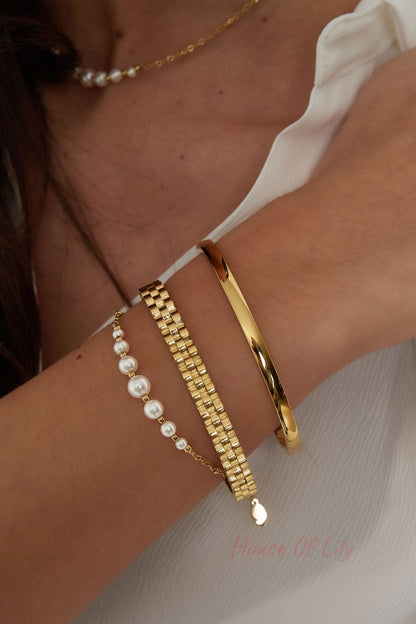 Model wrist wearing layered gold jewelry including a polished gold bangle, gold chain bracelets, and pearl chain necklace