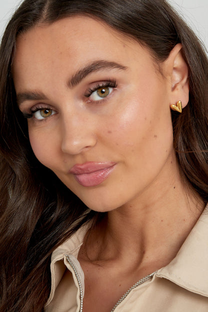Close-up of a woman wearing gold textured heart stud earrings, showing natural makeup and youthful style