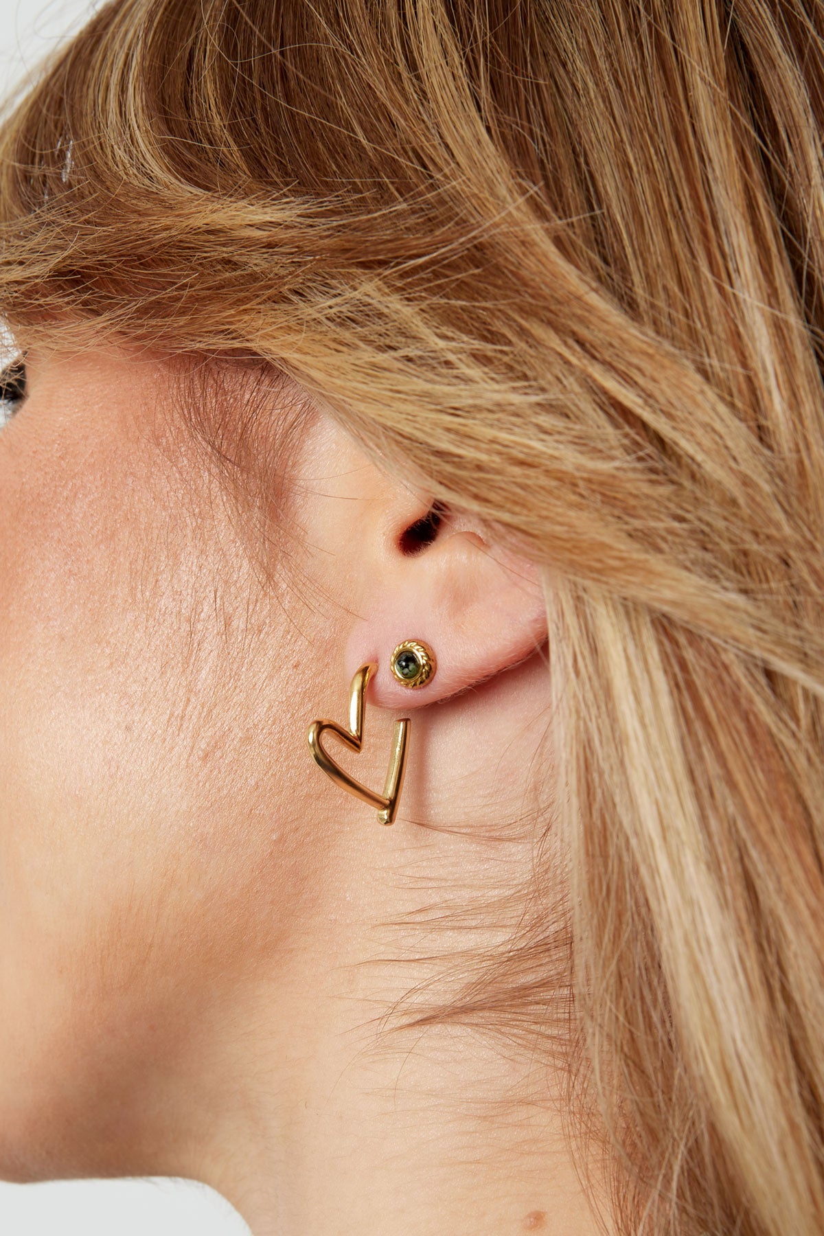 Close-up of woman's ear wearing gold heart hoop earrings with a small green stud earring