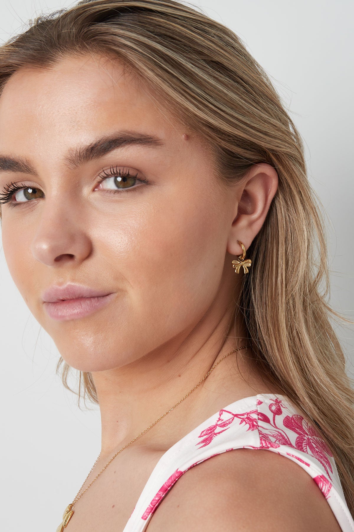 Close-up of a woman's face wearing one gold bow hoop earring, styled with a floral top and a delicate necklace.