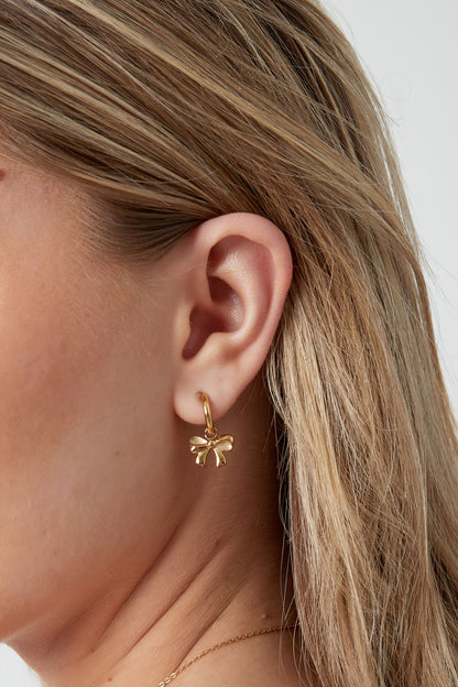 Side profile showing a woman's ear adorned with a small gold hoop earring featuring a dangling bow charm.