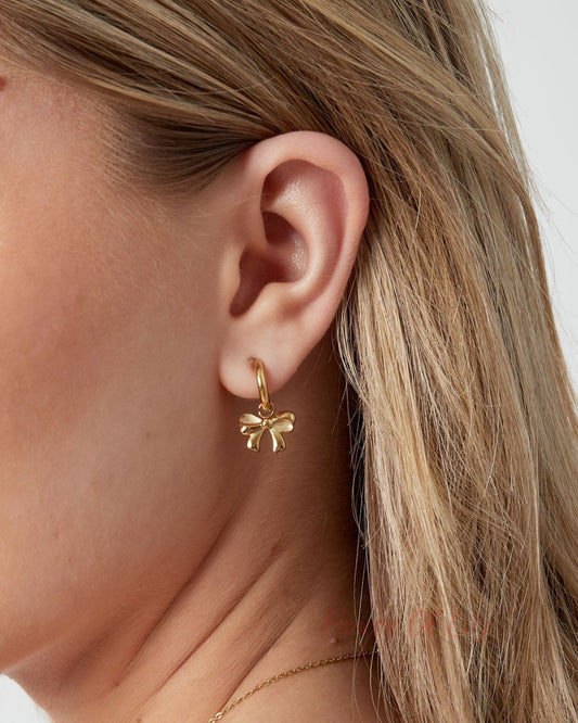 Side profile showing a woman's ear adorned with a small gold hoop earring featuring a dangling bow charm.