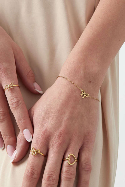 Model wearing gold bow charm bracelet and matching gold bow and heart rings with beige outfit