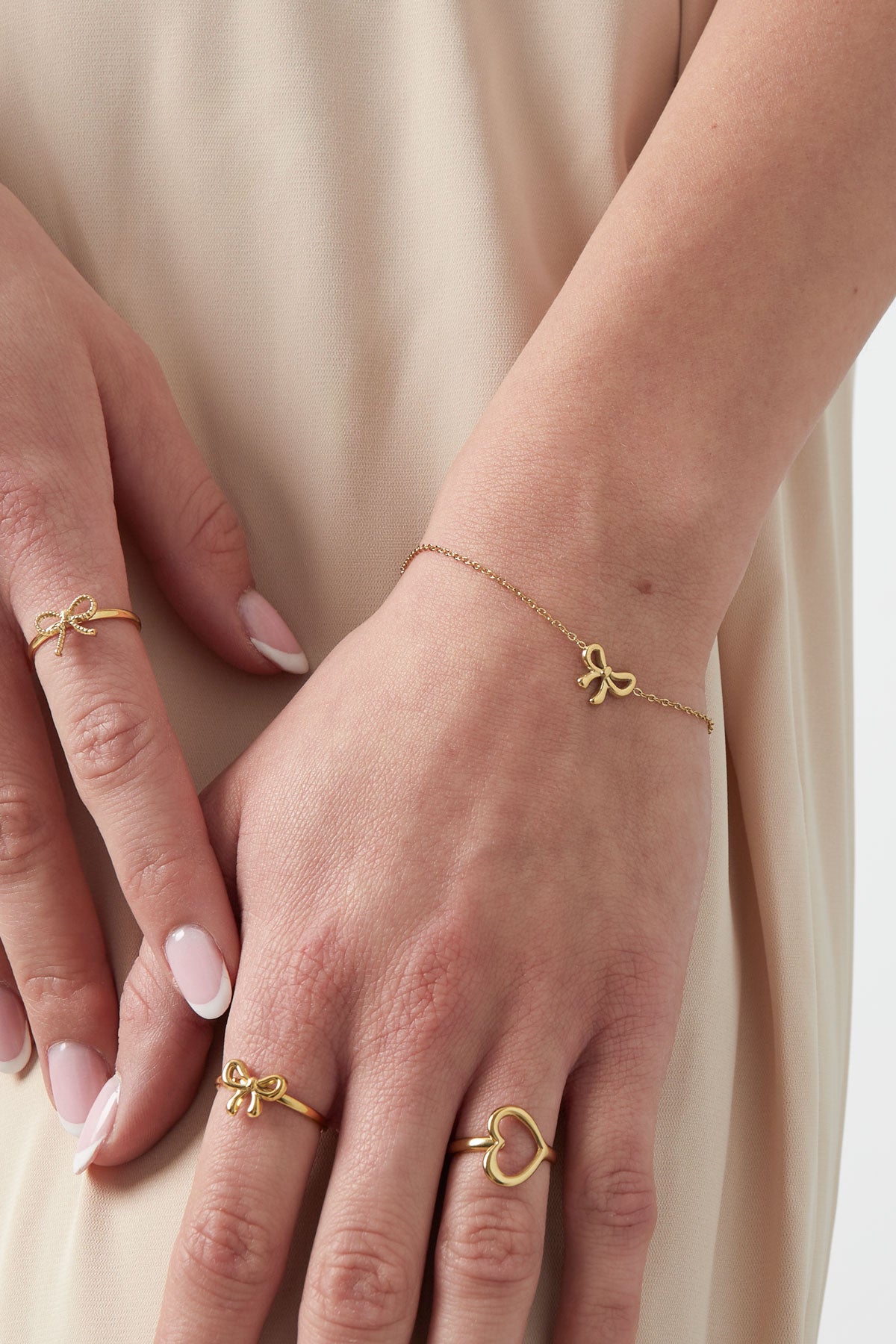 Model wearing gold bow charm bracelet and matching gold bow and heart rings with beige outfit
