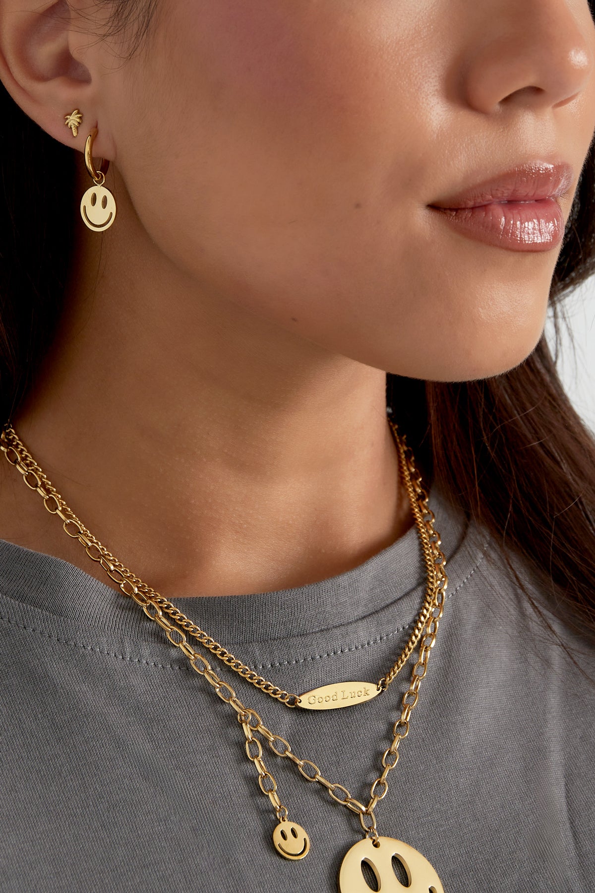 Close-up side view of a woman wearing gold smiley face hoop earrings paired with layered golden necklaces and a grey top