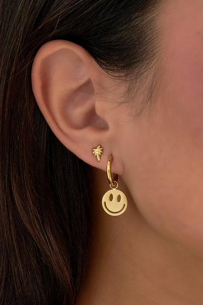 Profile close-up of a woman wearing a gold smiley face hoop earring alongside a small palm tree stud earring