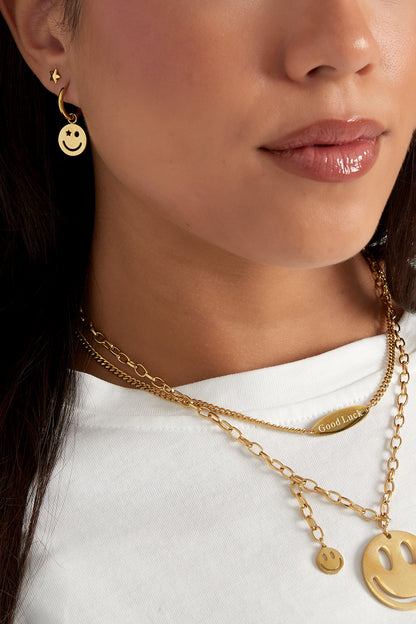 Close-up showing gold smiley face hoop earring worn with layered gold necklaces and star stud earring