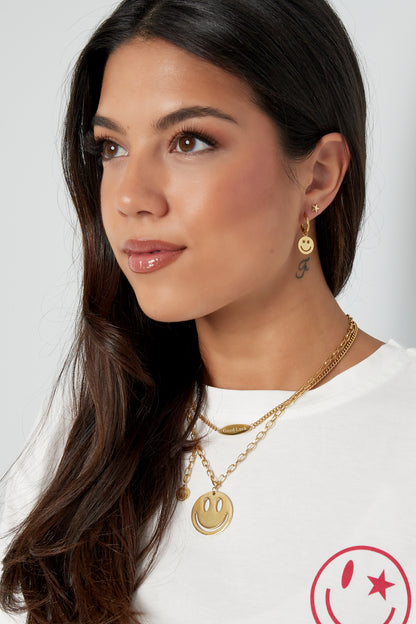 Portrait of woman smiling wearing gold smiley face hoop earrings and layered pendant necklaces