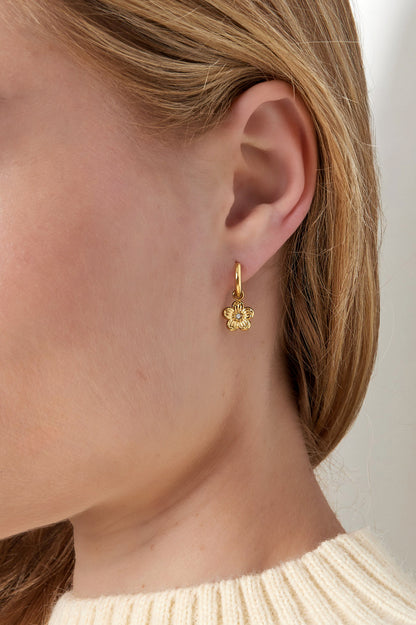 Close up of a woman wearing gold hoop earrings with dangling flower charms and rhinestone centres, styled with a cream knitted sweater