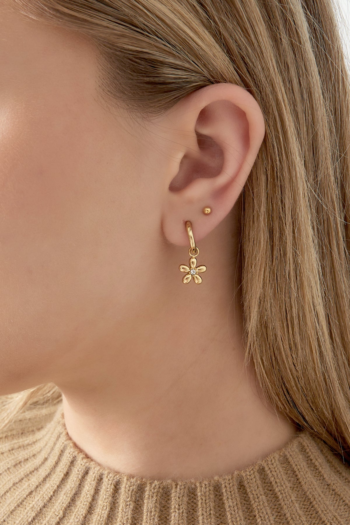 Close-up of a woman's ear wearing a single gold drop earring with a flower charm accented with rhinestone, styled with light brown hair and a beige knit top