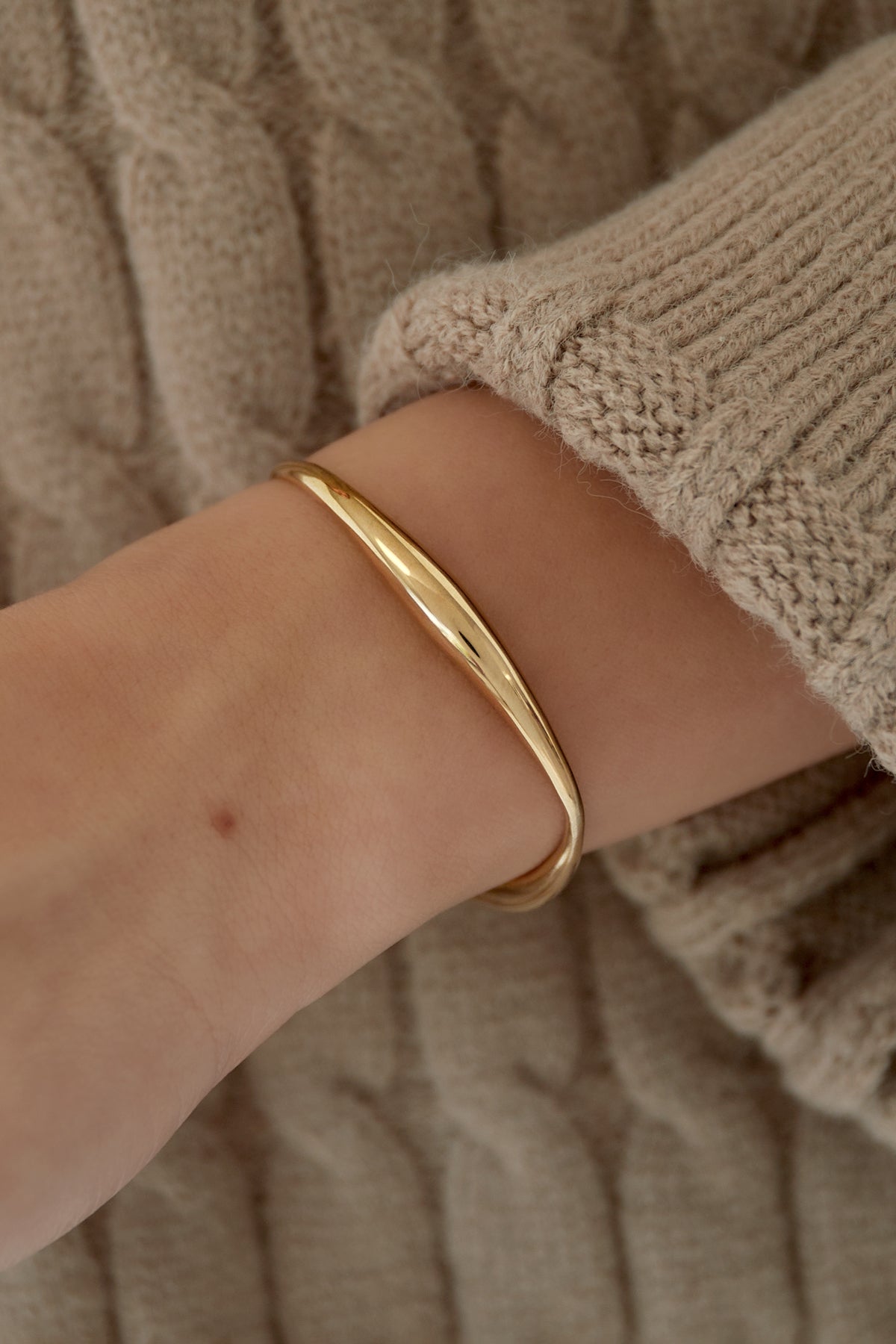 Close-up of gold cuff bracelet worn on wrist with beige knitted sweater, showcasing polished surface and soft design