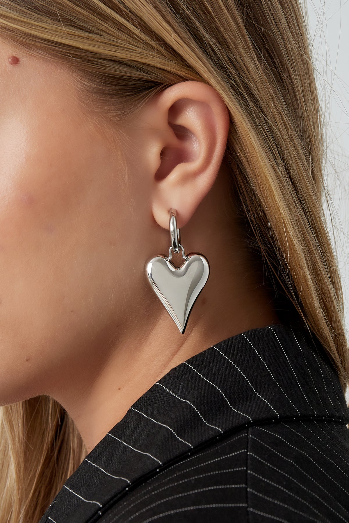 Close-up of a blonde woman wearing silver chubby heart-shaped hoop earrings paired with a black pinstriped jacket