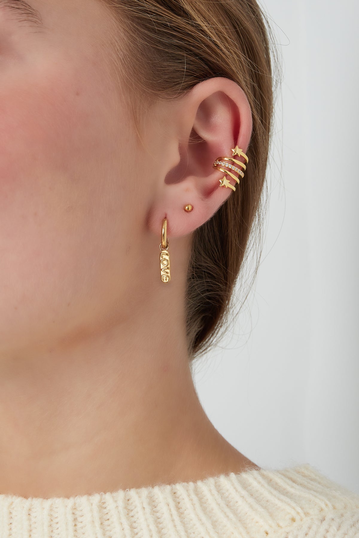 Gold-tone spiral star ear cuff fitted on upper ear alongside small gold studs and a hanging love charm earring