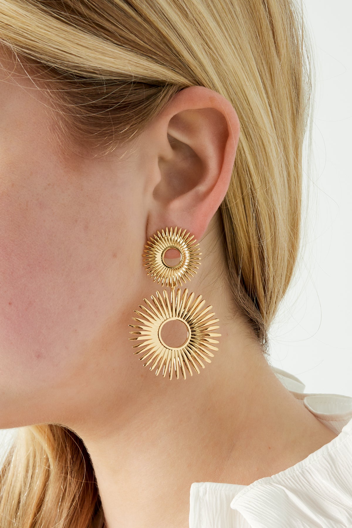 Close-up of radiant gold sunburst earrings worn by woman with blonde hair and white blouse