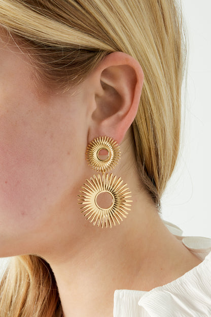 Close-up of radiant gold sunburst earrings worn by woman with blonde hair and white blouse