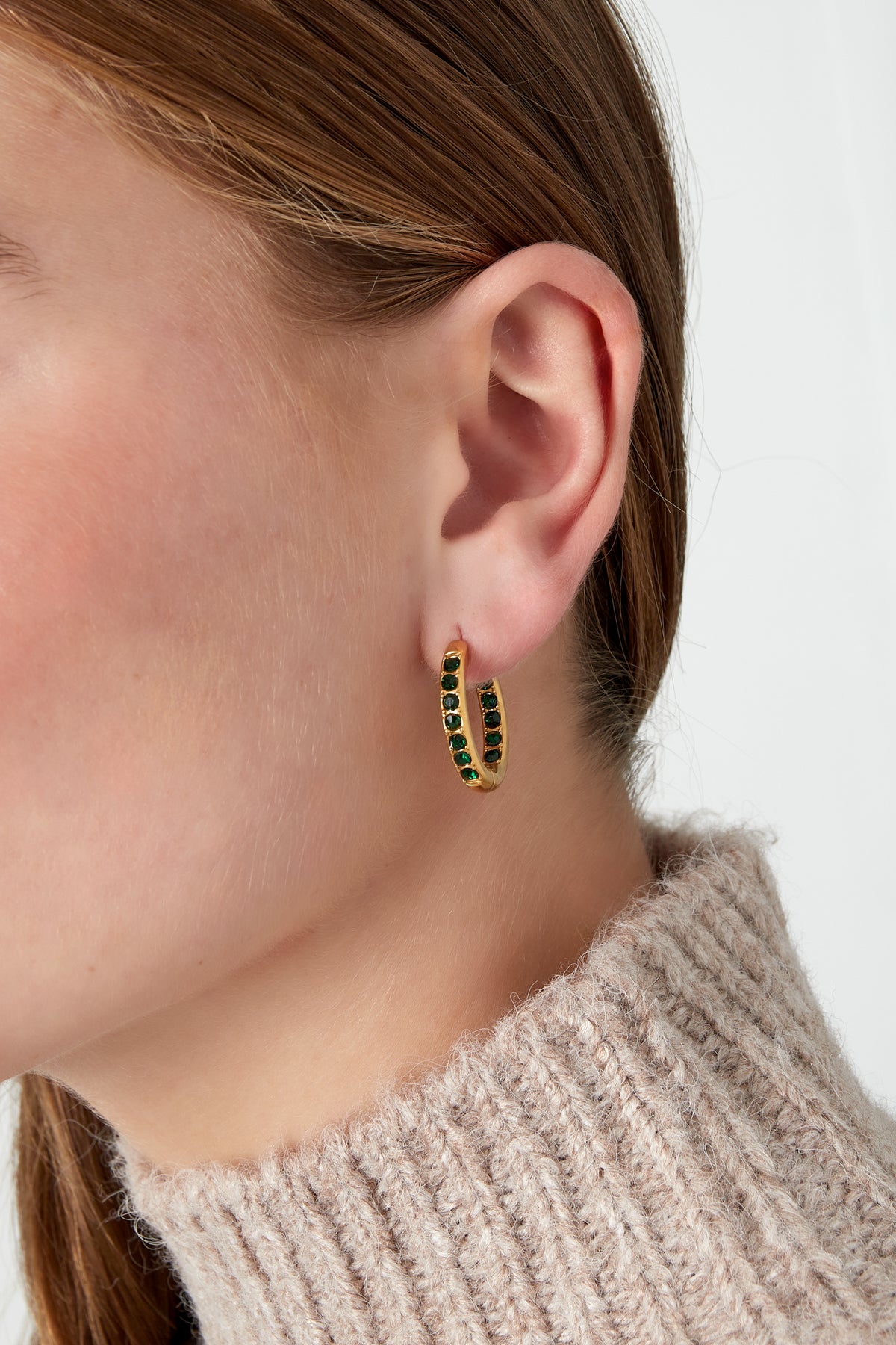 Close-up of a woman wearing gold hoop earrings with green rhinestones, styled with a cozy knit sweater
