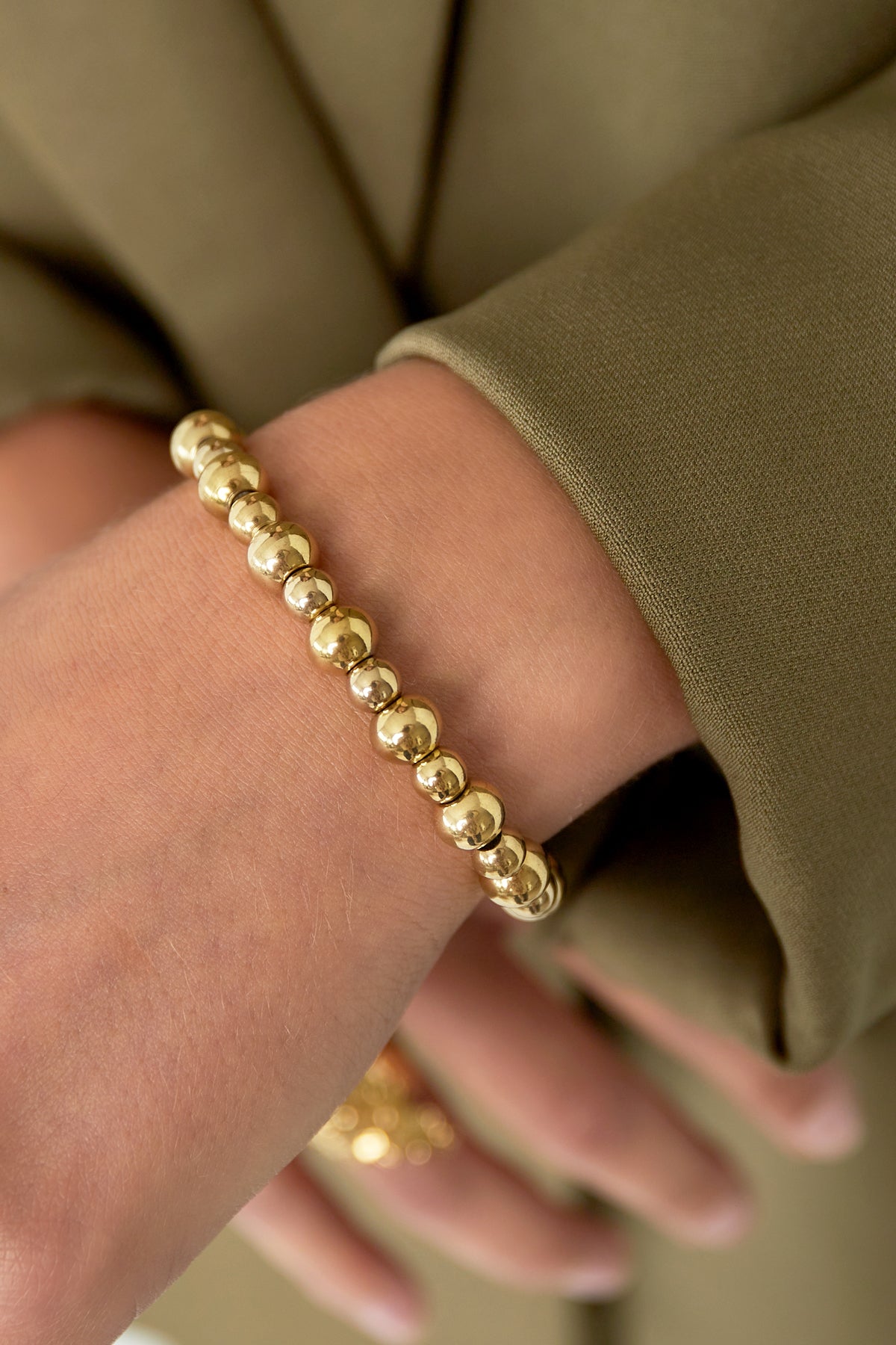 Close-up of wrist wearing golden beaded bracelet with smooth polished beads, styled with olive green sleeve