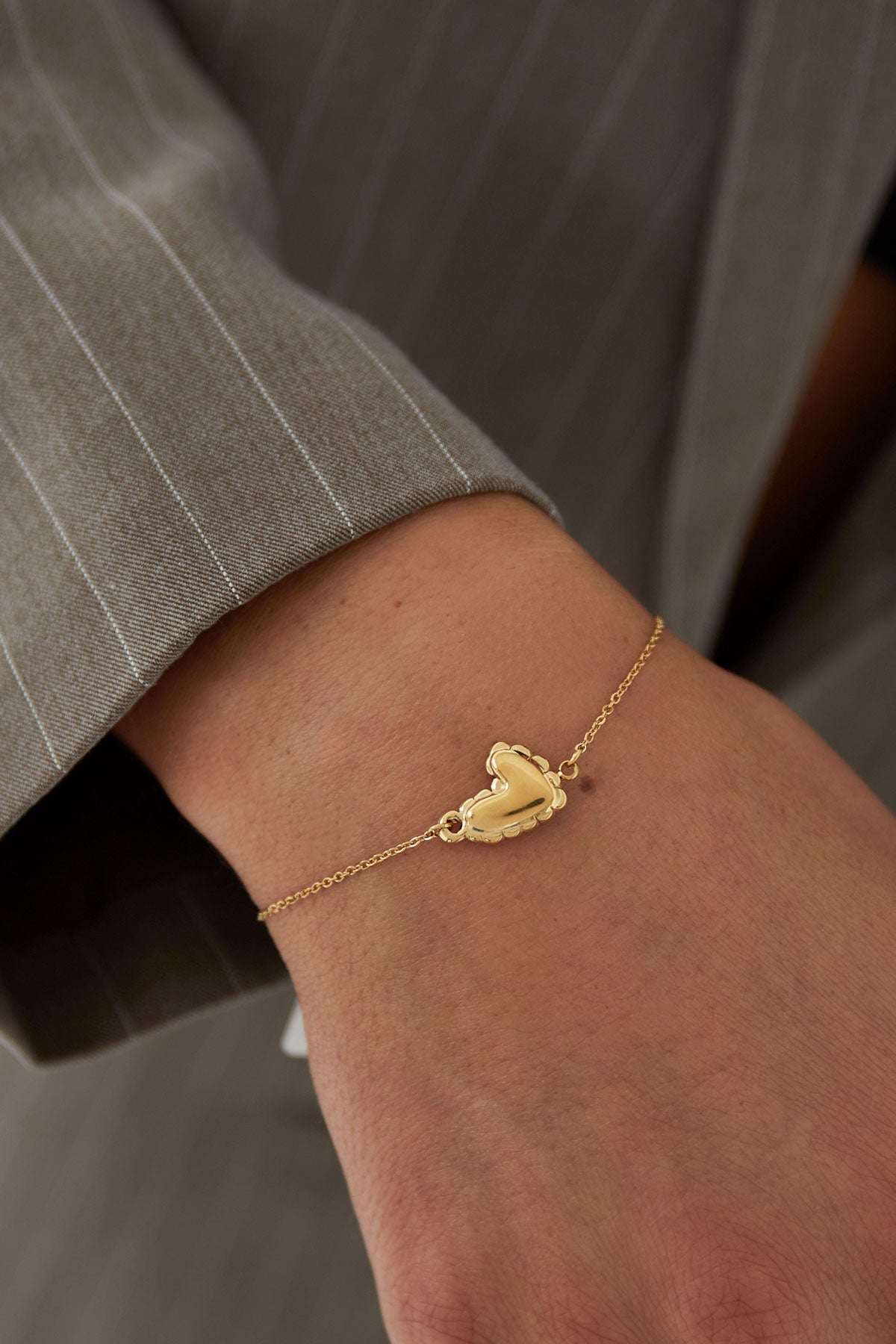 Close-up of gold heart bracelet worn on wrist with grey pinstripe sleeve visible