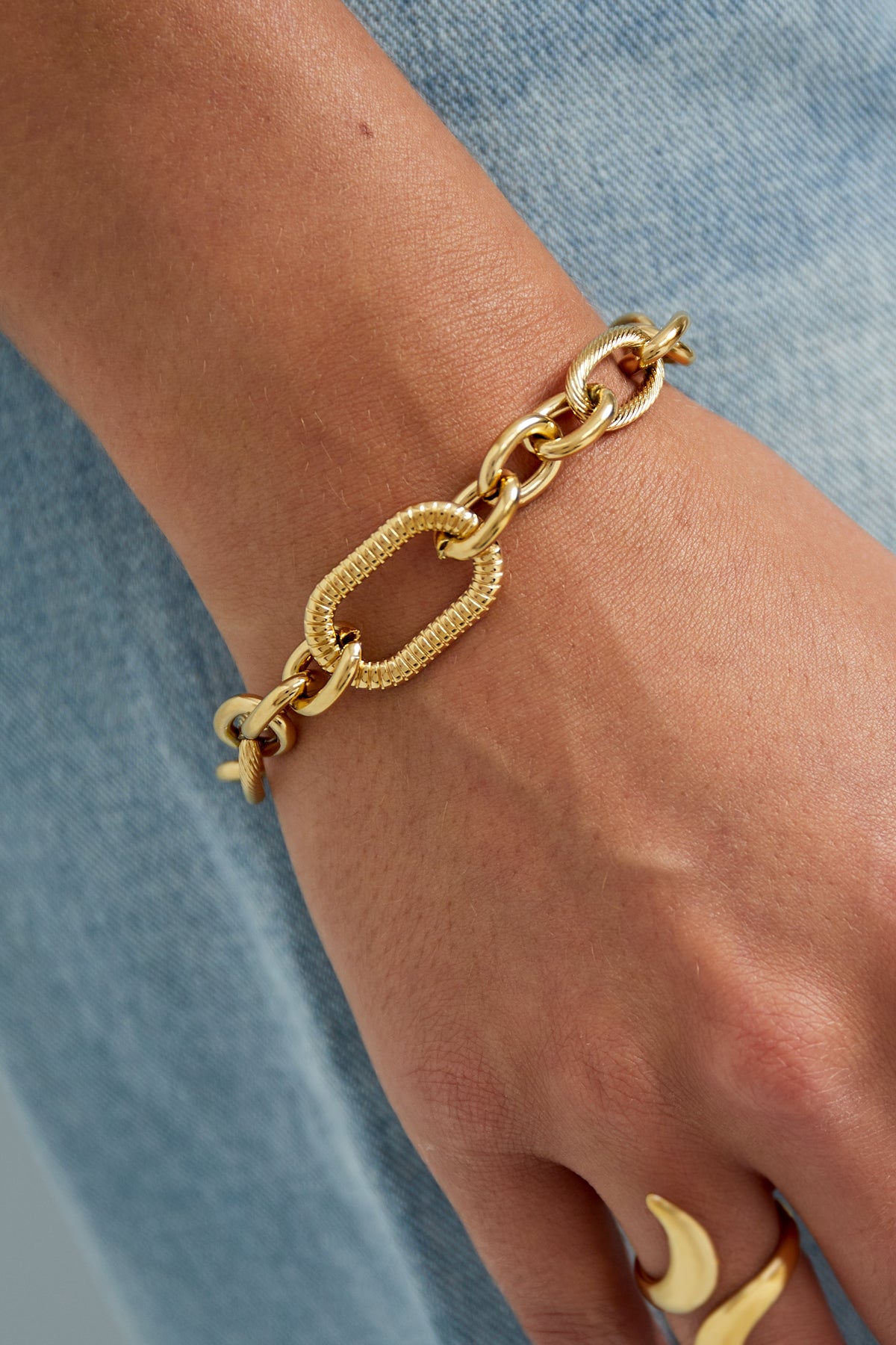 Close-up of gold textured link chain bracelet worn on wrist paired with gold rings and denim