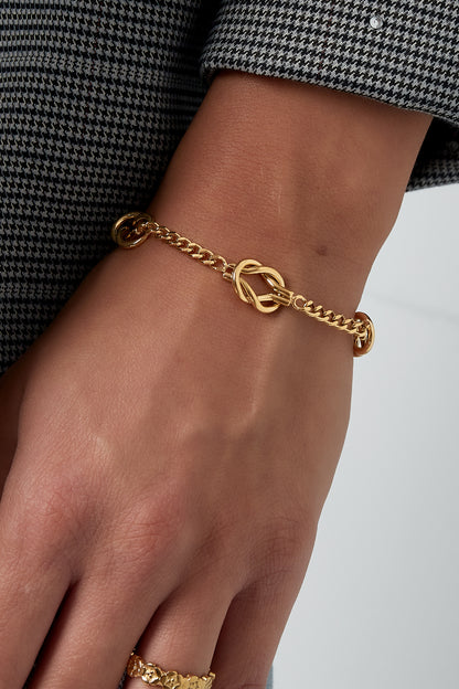 Model's wrist wearing a delicate gold bracelet with three intertwined knot details, styled with a houndstooth sleeve