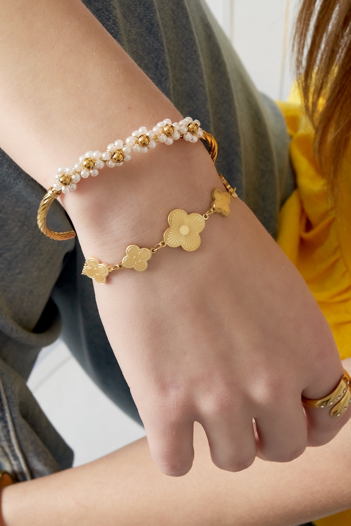 Woman's wrist wearing the gold-tone pearl flower cuff bracelet paired with another delicate gold flower bracelet and a gold ring, styled with casual outfit.