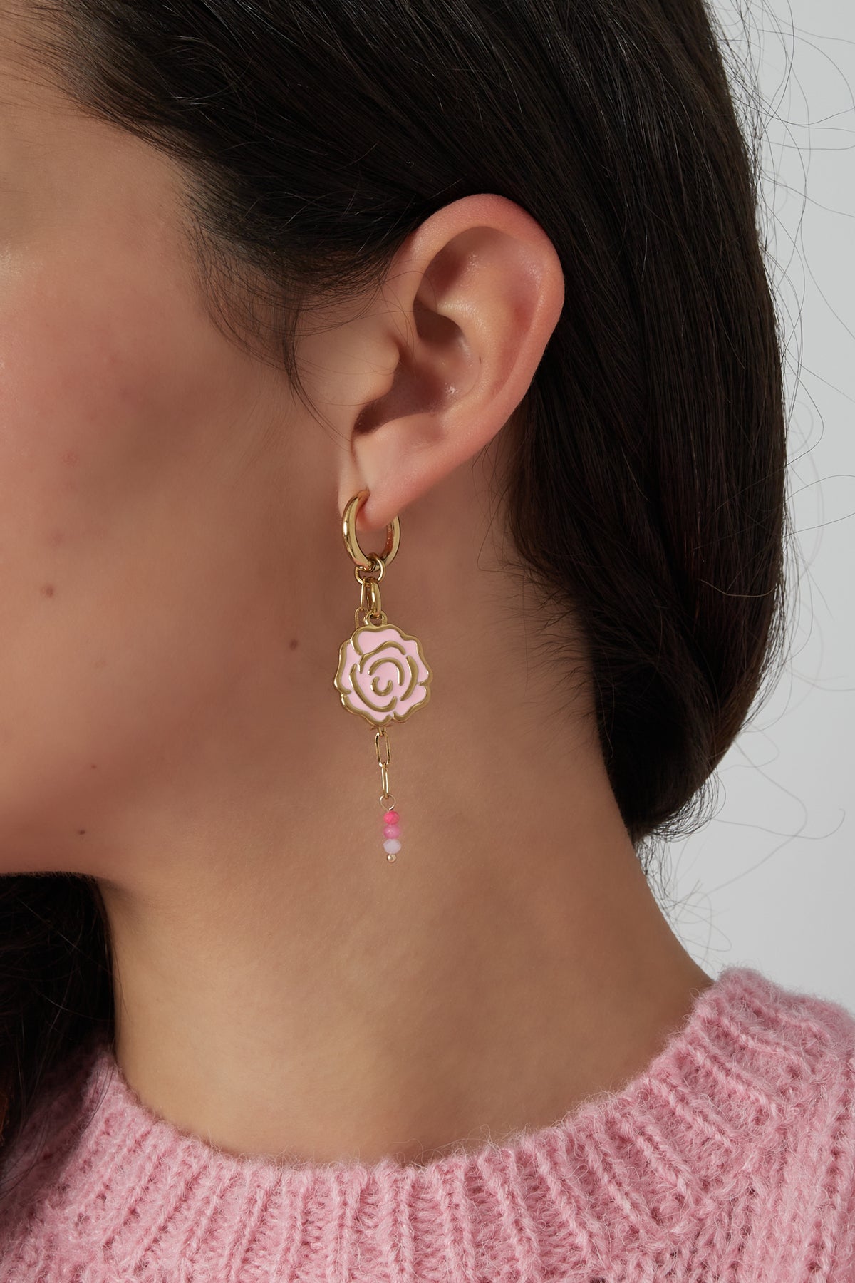 Close-up of woman wearing pink enamel rose drop earring hanging from a gold-tone hoop with pink glass beads, paired with a pink knit sweater