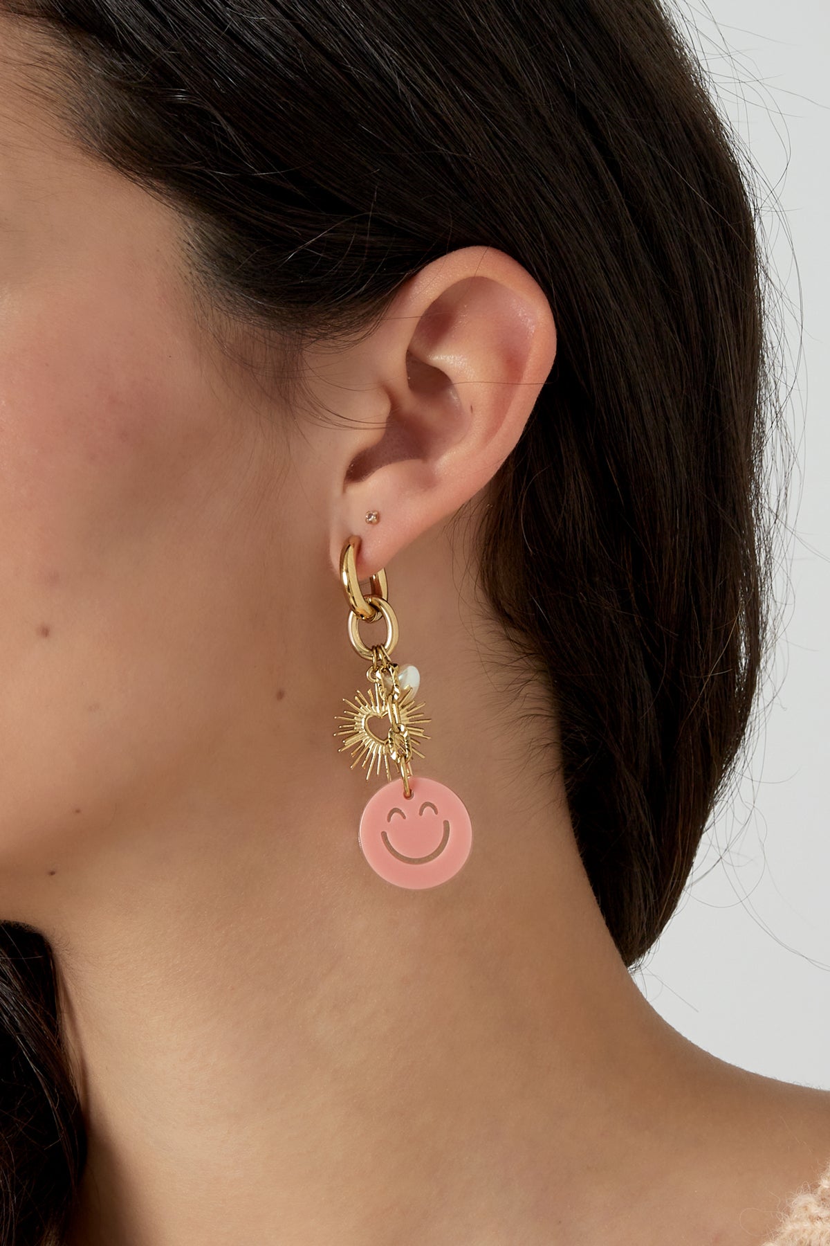 Side profile of woman wearing the gold drop earring with pink smiley charm and sunburst heart, showcasing stylish youthful look
