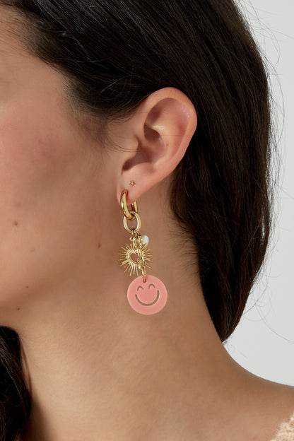 Side profile of woman wearing the gold drop earring with pink smiley charm and sunburst heart, showcasing stylish youthful look