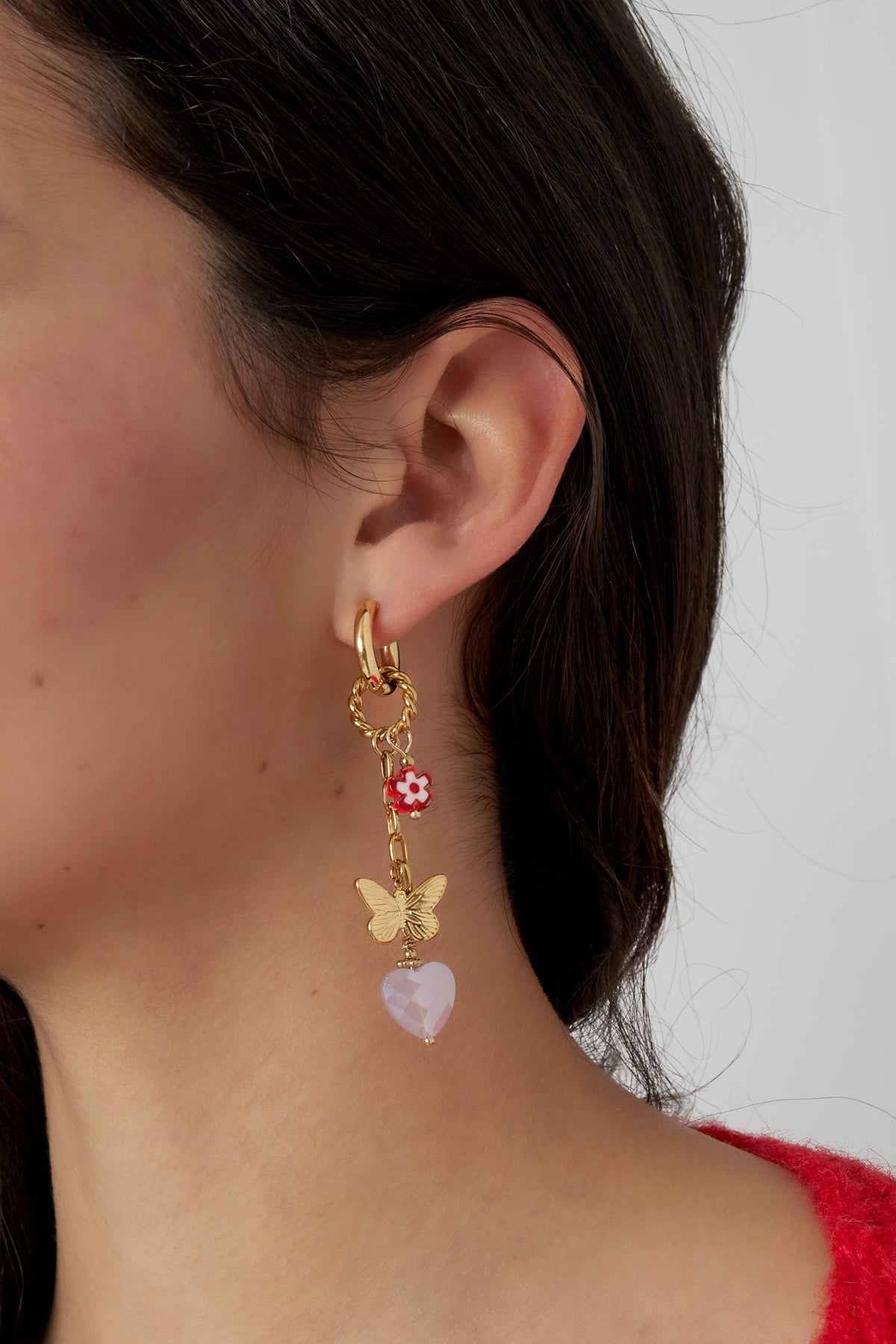 Woman wearing gold drop earrings with red flower, butterfly charm, and faceted pink heart pendant