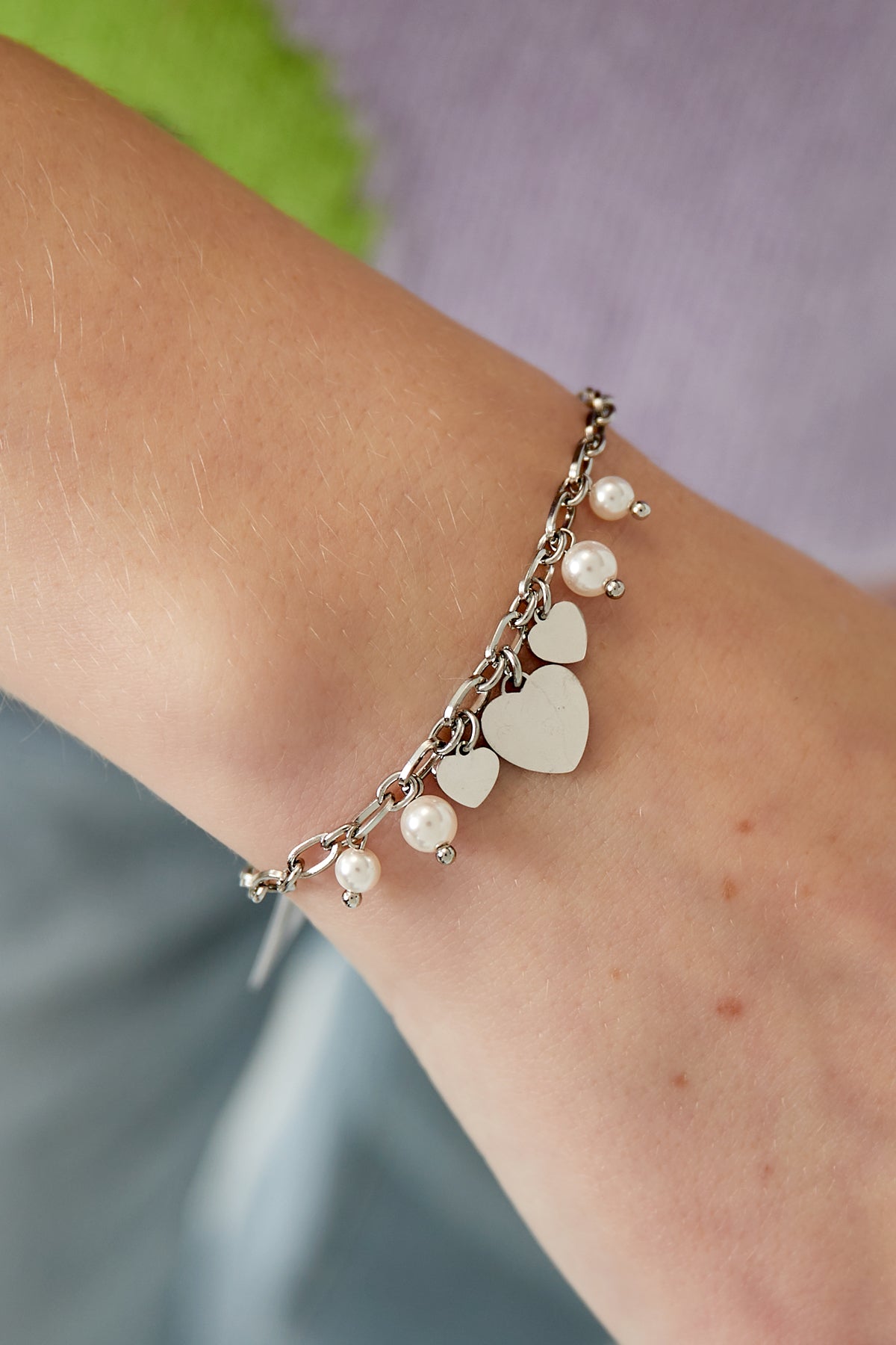 Silver-tone charm bracelet with heart pendants and pearl beads worn on wrist against colorful background.