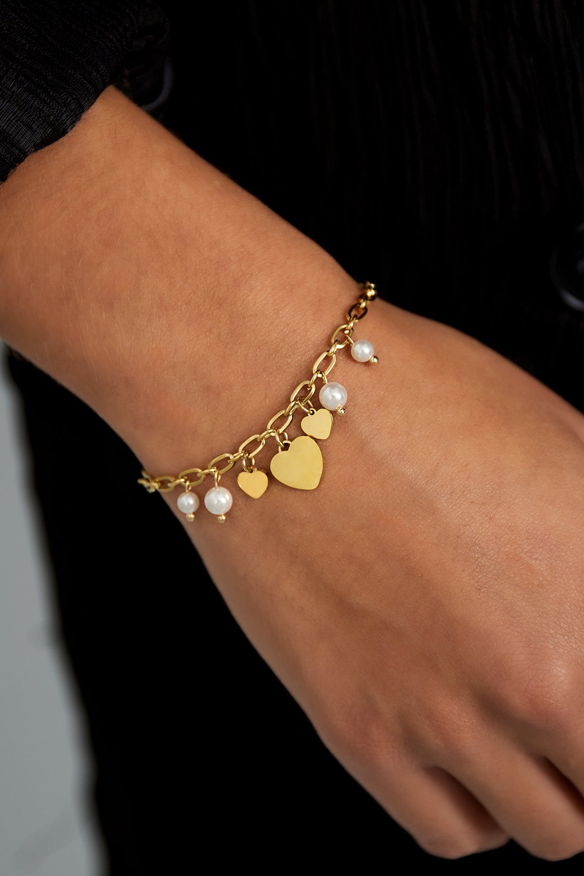 Close-up of gold-tone hearts and pearls bracelet worn on wrist, highlighting soft aesthetic and delicate design.