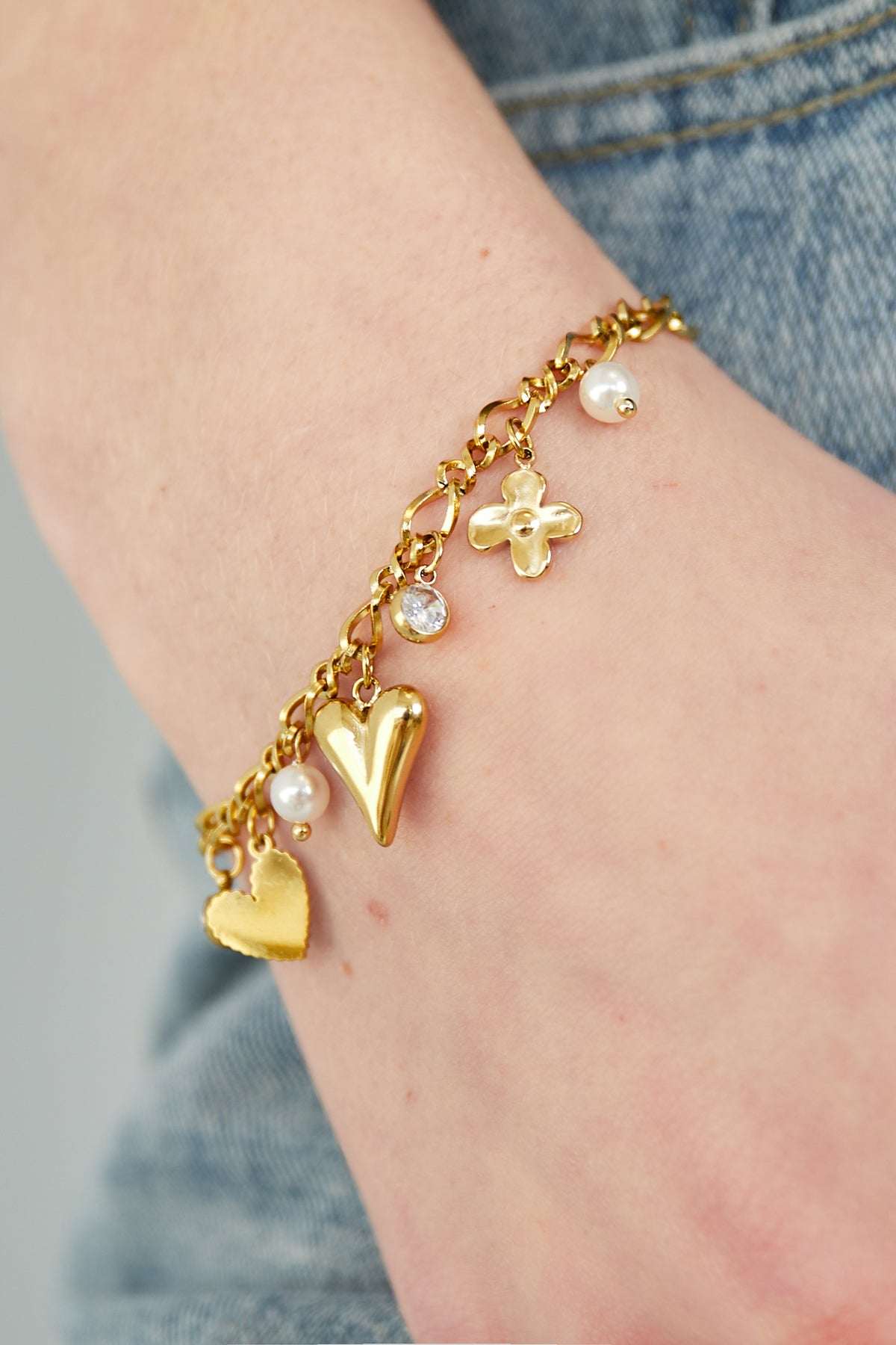 Close-up of the gold charm bracelet worn on wrist, showing detailed heart, flower, rhinestone and pearl charms paired with denim
