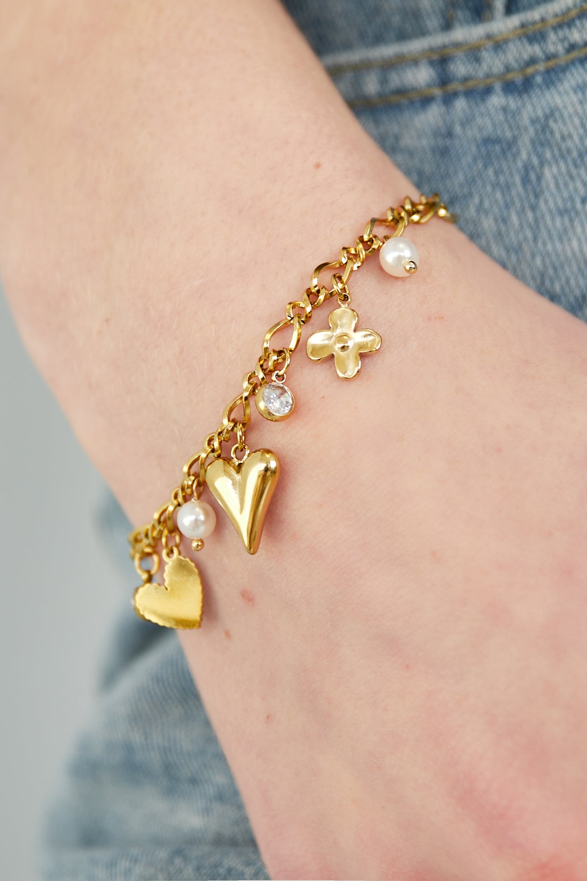 Close-up of the gold charm bracelet worn on wrist, showing detailed heart, flower, rhinestone and pearl charms paired with denim