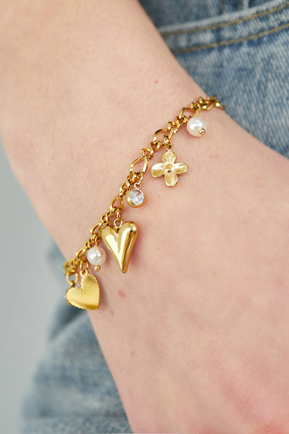 Close-up of the gold charm bracelet worn on wrist, showing detailed heart, flower, rhinestone and pearl charms paired with denim