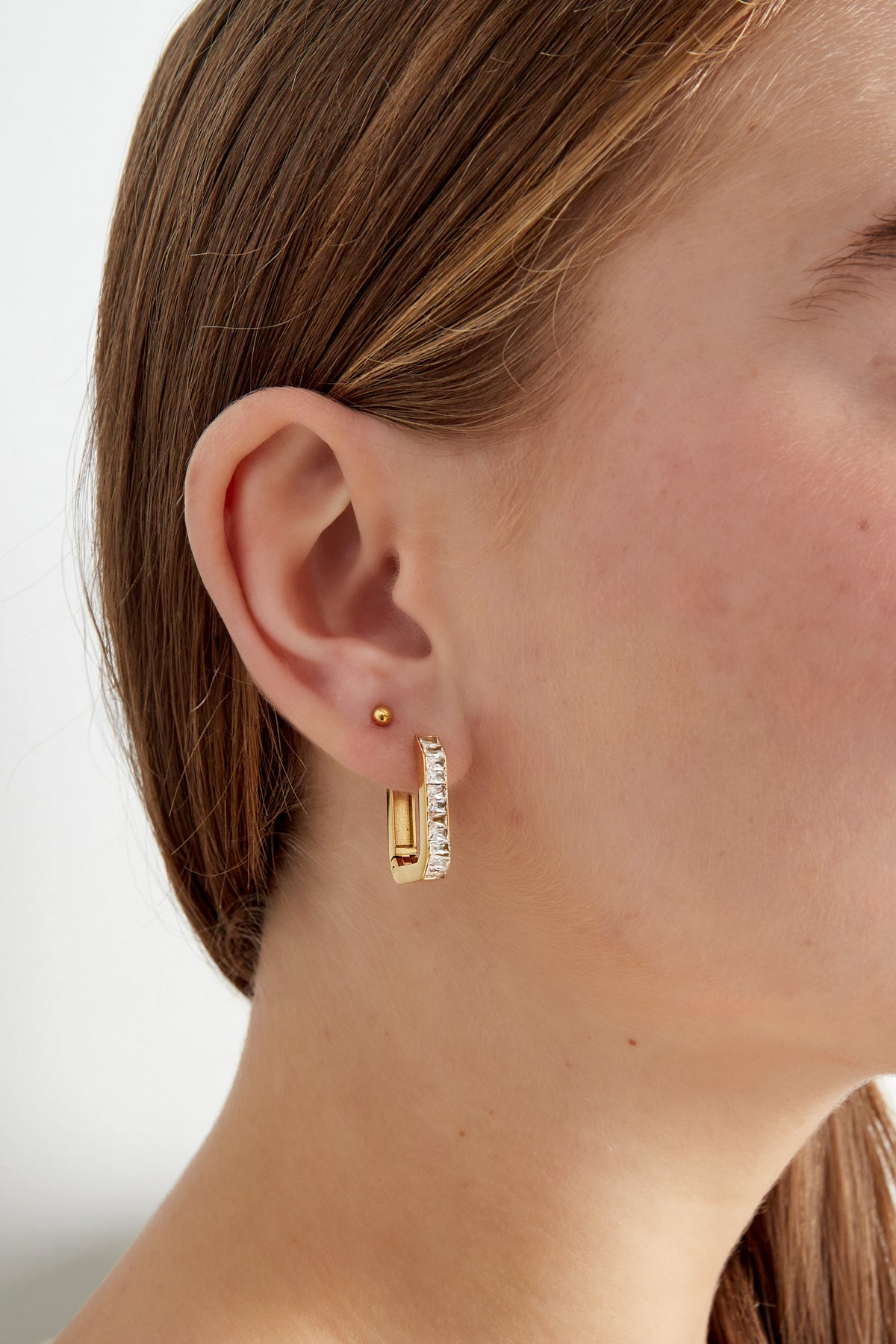 Close-up of model wearing gold rectangular crystal hoop earring on ear with small gold stud