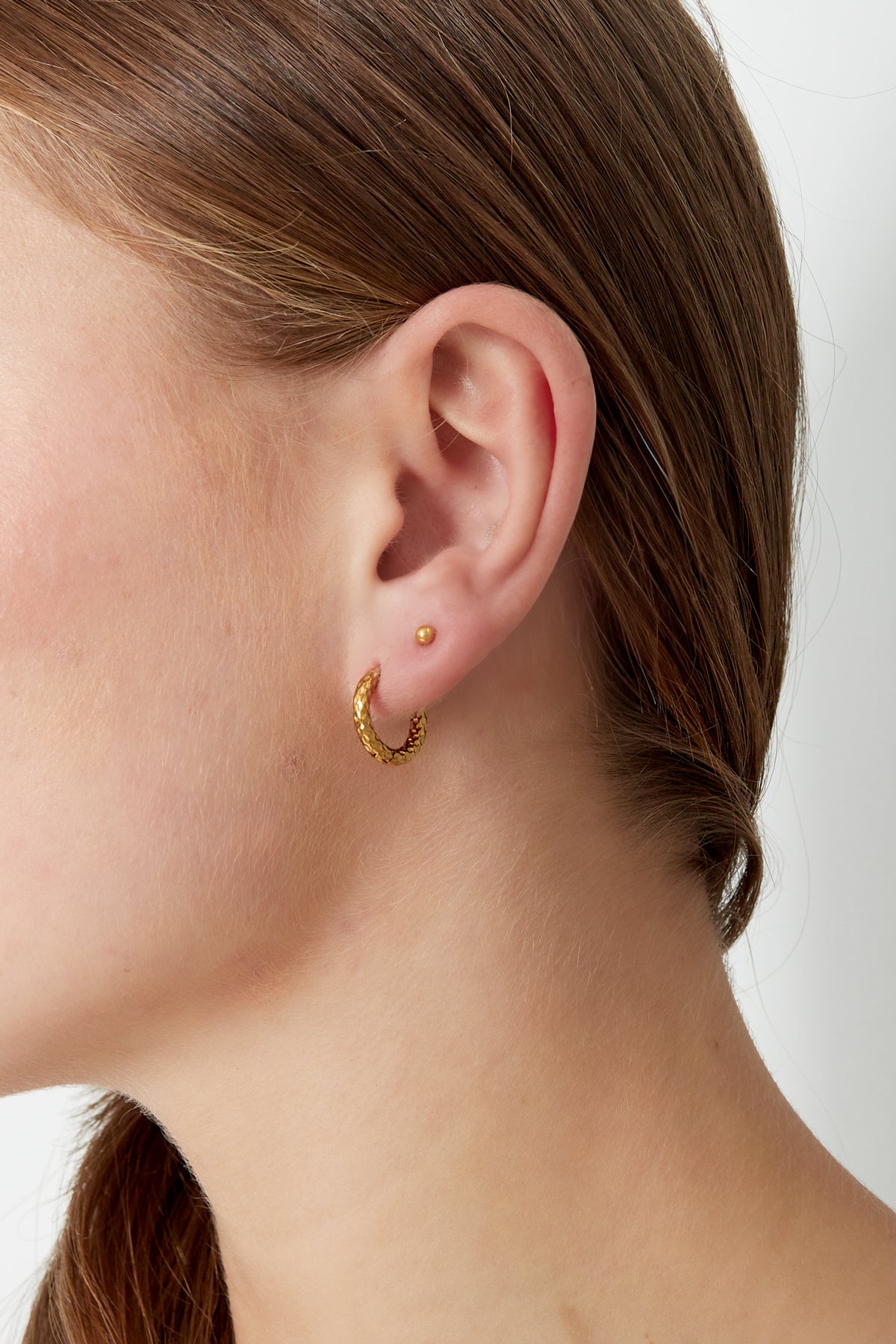 Close-up of a woman's ear wearing textured gold hoop earring, highlighting its youthful and minimal aesthetic
