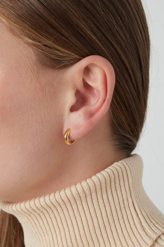Close-up of gold mini hoop earring worn by woman with light skin and brown hair wearing cream turtleneck