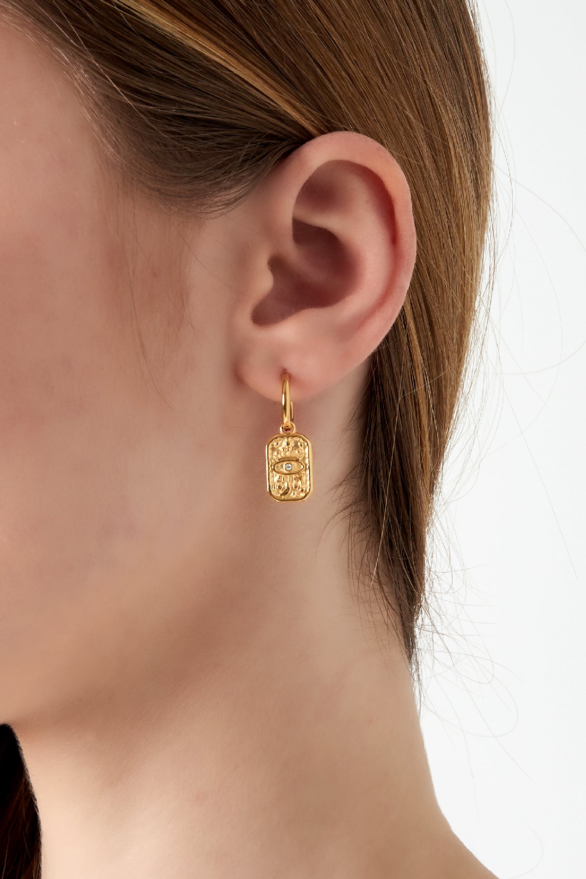 Close-up of a woman's ear wearing a gold hoop earring with a rectangular mystic eye charm pendant