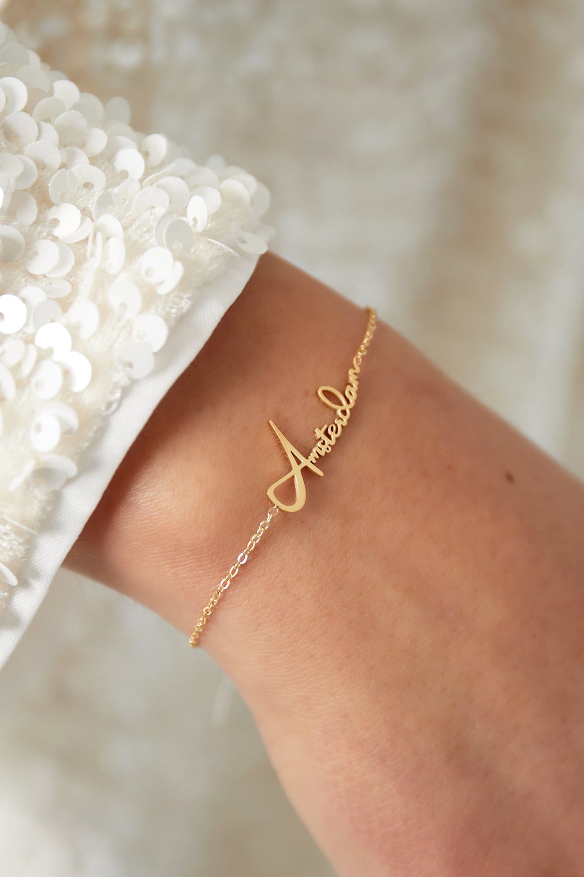 Close-up of gold Amsterdam Script Bracelet worn on wrist with white sequined sleeve