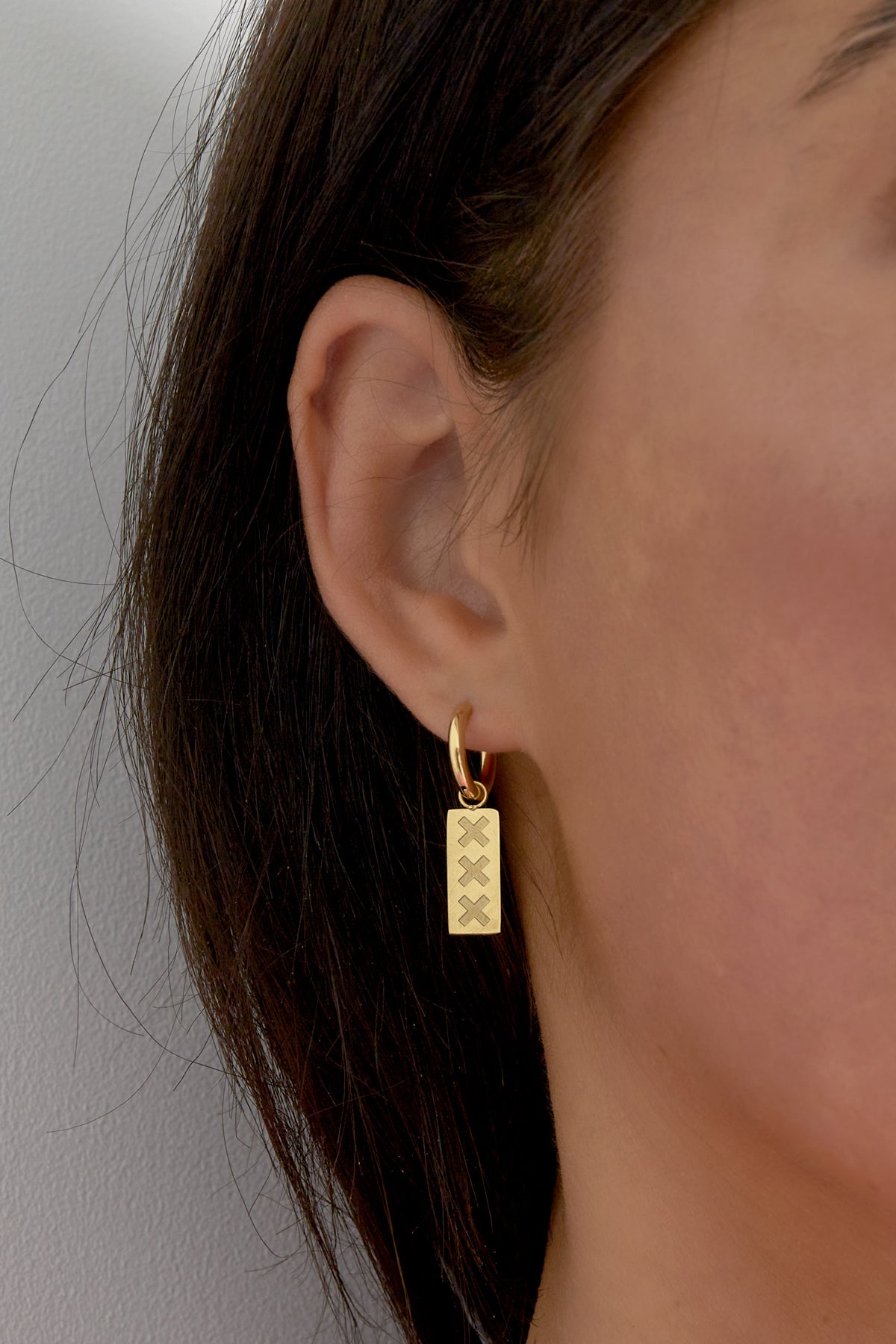 Close-up of woman wearing gold Amsterdam minimalist drop earring with triple X engraved rectangular pendant